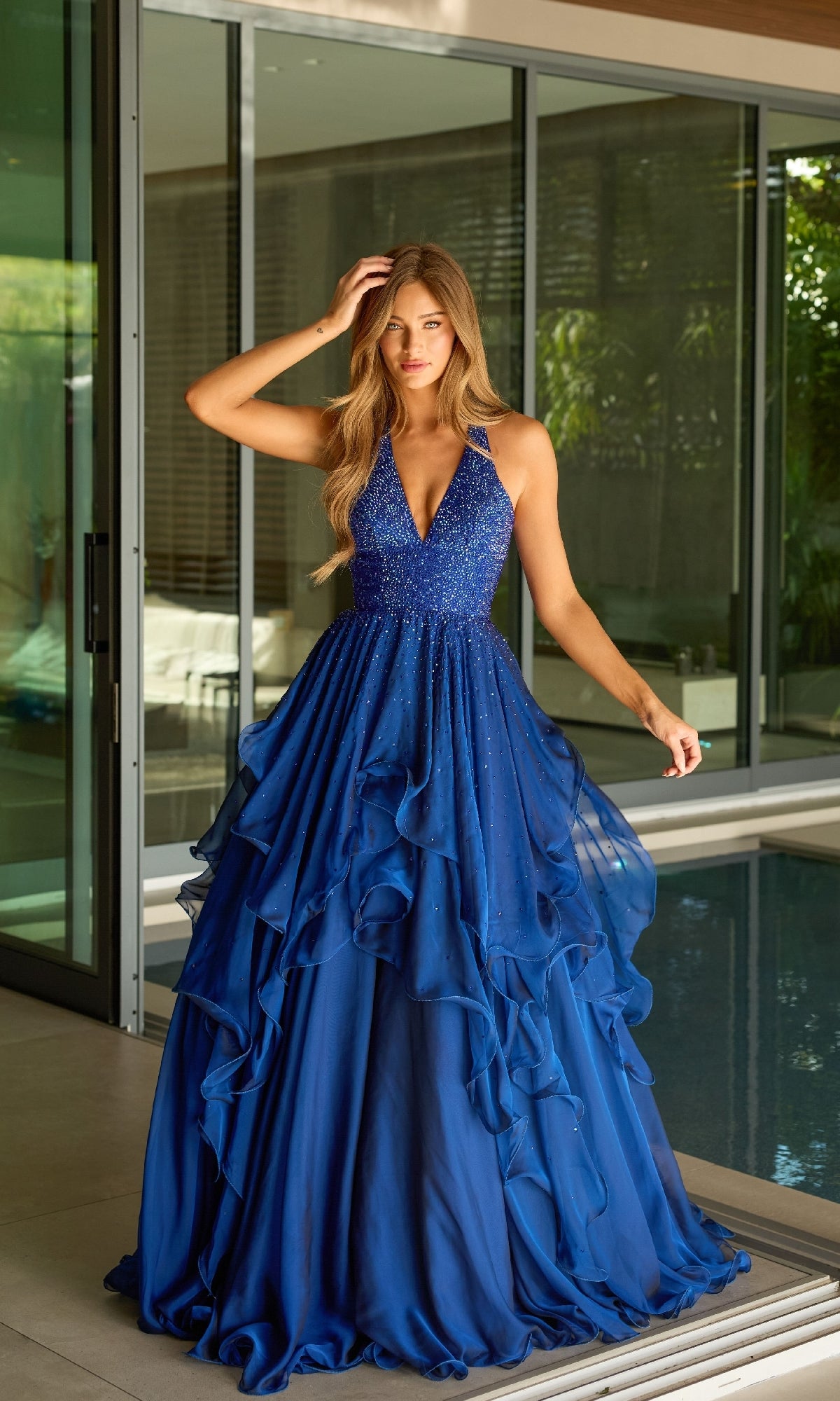 Indoors by a pool, a woman wears the Ava Presley 42235 Long Halter A-Line Formal Dress in deep blue, featuring a sleeveless design with ruffles, a sparkling bodice, and a layered skirt.
